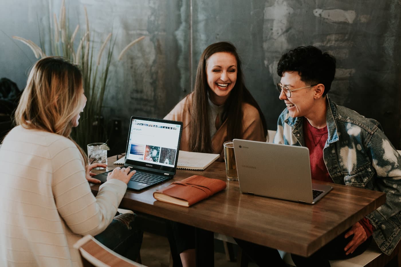 Three women sit at a desk with their laptops. They are chatting and laughing. (Alle lachen)